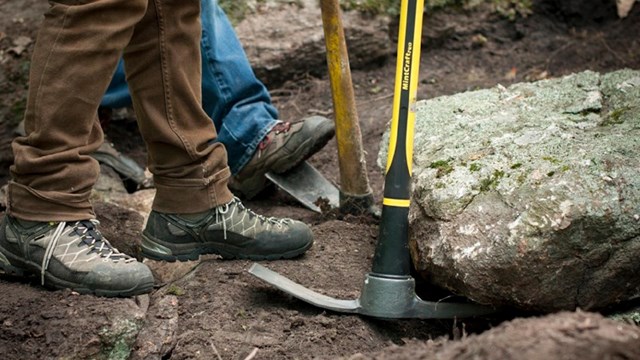 Close up of two people working with picks to move a large rock on the trail.